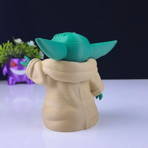 Peut inclure: Figurine imprim&eacute;e en 3D d'un personnage aux grandes oreilles et &agrave; la peau verte, v&ecirc;tu d'une robe beige. La figurine est pos&eacute;e avec un bras lev&eacute;. Le fond est sombre, et un pot violet avec des fleurs blanches est visible.