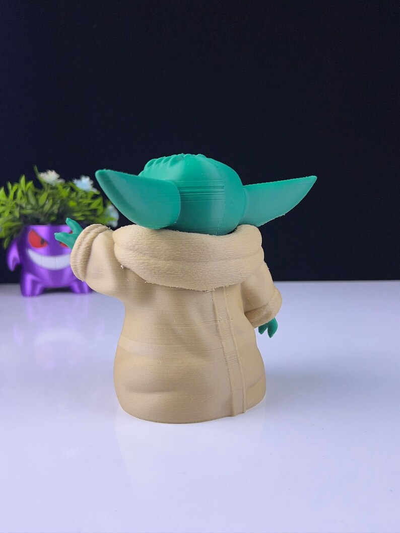 Peut inclure: Figurine imprim&eacute;e en 3D d'un personnage aux grandes oreilles et &agrave; la peau verte, v&ecirc;tu d'une robe beige. La figurine est pos&eacute;e avec un bras lev&eacute;. Le fond est sombre, et un pot violet avec des fleurs blanches est visible.