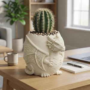 May include: A cream-colored dragon-shaped planter holding a green cactus. The planter has detailed dragon wings and a head sculpted on the front. The cactus is planted in a bed of small pebbles. The planter is on a wooden table.