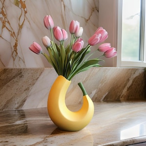 Puede incluir: Un jarrón de cerámica amarilla con forma de plátano sostiene un ramo de tulipanes rosas con tallos y hojas verdes. El jarrón tiene un diseño curvo con un detalle de tallo verde. El arreglo está sobre una encimera de mármol.
