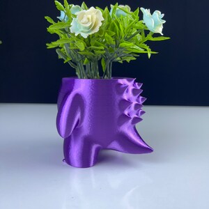 Pu&ograve; includere: Un vaso viola, stampato in 3D, a forma di testa di dinosauro stilizzata. Il vaso contiene fiori artificiali con rose bianche e azzurre e foglie verdi. Il vaso &egrave; su una superficie bianca.