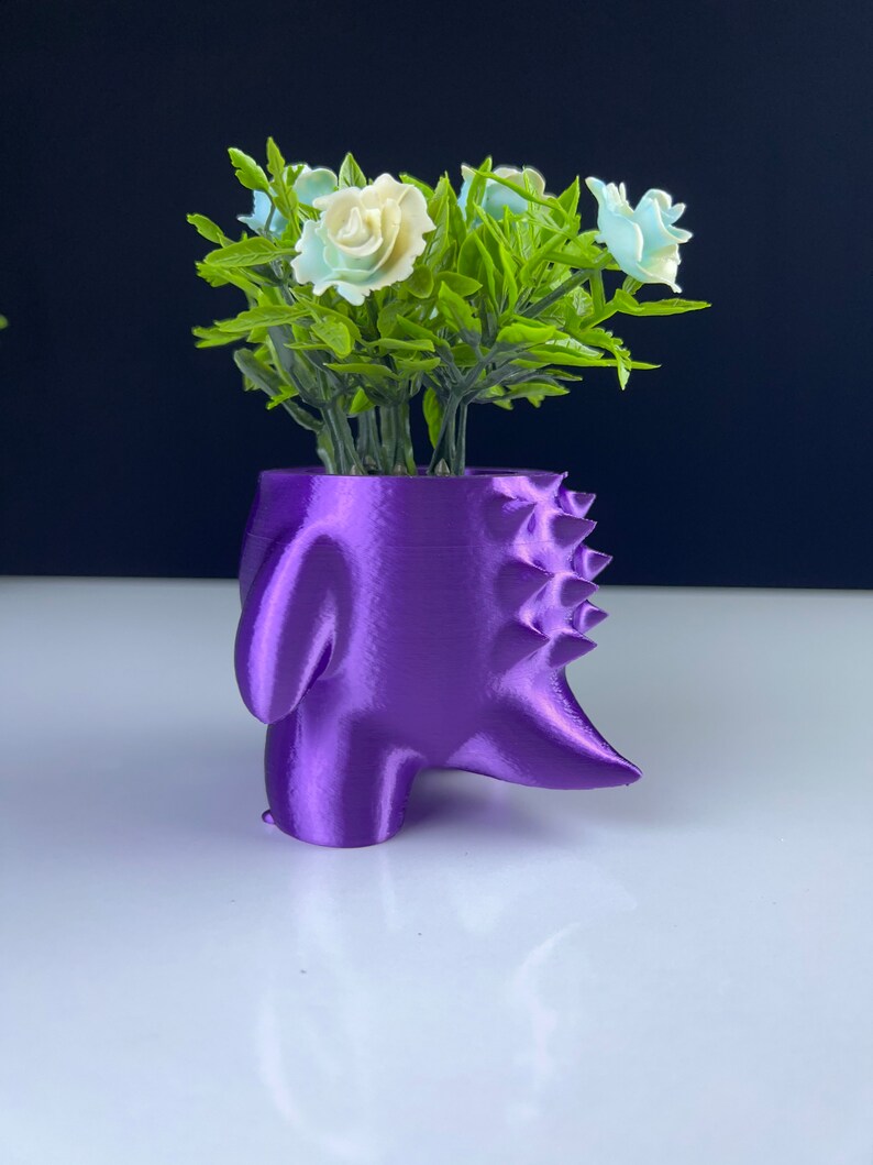 Pu&ograve; includere: Un vaso viola, stampato in 3D, a forma di testa di dinosauro stilizzata. Il vaso contiene fiori artificiali con rose bianche e azzurre e foglie verdi. Il vaso &egrave; su una superficie bianca.