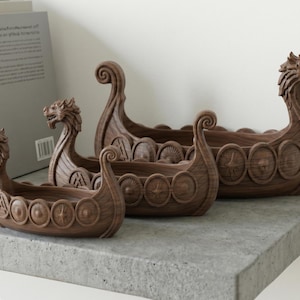 Könnte beinhalten: Drei braune dekorative Schalen aus Holz in Form von Wikingerschiffen. Jedes Schiff hat einen Drachenkopf am Bug und Schilde an den Seiten. Die Schalen sind in einer Reihe angeordnet, unterschiedlich groß und für die Inneneinrichtung geeignet.