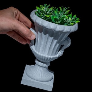 Può includere: Un vaso grigio, ornato a forma di urna, con una base quadrata, che contiene un vivace fogliame verde. Il vaso ha una superficie dettagliata e strutturata e manici decorativi. L'immagine è su sfondo nero.