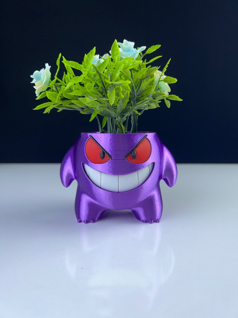 Pu&ograve; includere: Un vaso viola a forma di personaggio dei cartoni animati, con occhi rossi e un ampio sorriso, contenente fogliame verde e fiori azzurri. Il vaso ha una superficie strutturata ed &egrave; posto su uno sfondo nero.