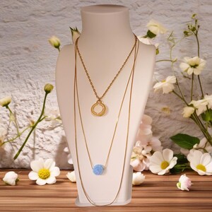 May include: Three gold necklaces displayed on a white mannequin. The top necklace has a circular pendant with text, the middle necklace has a light blue bead, and the third is a long chain. White flowers and greenery are in the background.