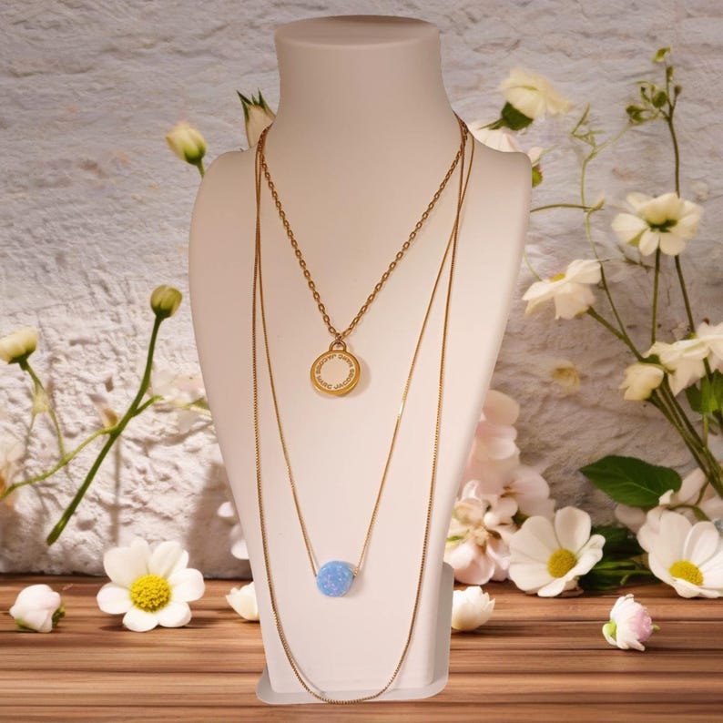 May include: Three gold necklaces displayed on a white mannequin. The top necklace has a circular pendant with text, the middle necklace has a light blue bead, and the third is a long chain. White flowers and greenery are in the background.