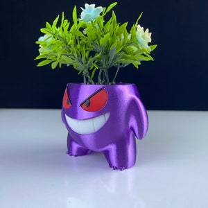 Puede incluir: Una maceta morada impresa en 3D con forma de personaje de dibujos animados con una amplia sonrisa y ojos rojos. La maceta contiene follaje verde artificial con flores azul claro. La maceta está sobre una superficie blanca con un fondo oscuro.