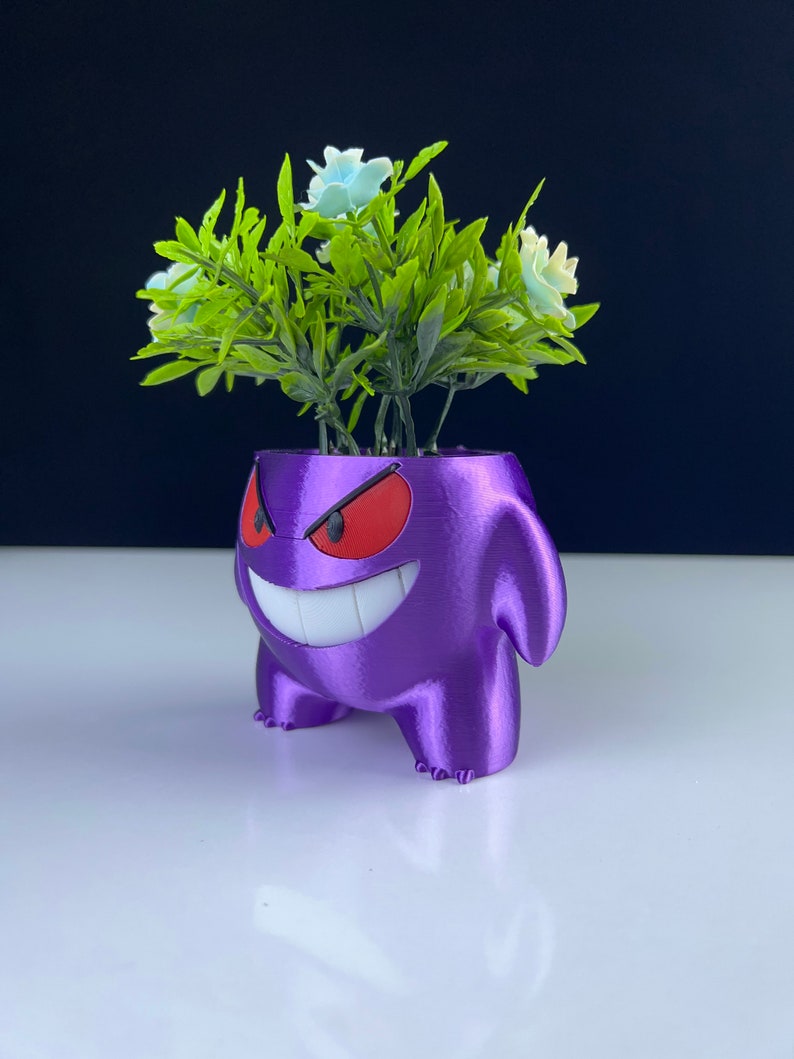 Pu&ograve; includere: Un vaso viola stampato in 3D a forma di personaggio dei cartoni animati con un ampio sorriso e occhi rossi. Il vaso contiene fogliame verde artificiale con fiori azzurri. Il vaso &egrave; su una superficie bianca con uno sfondo scuro.