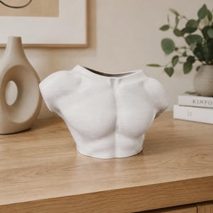 Puede incluir: Un jarrón de cerámica blanca con forma de torso humano, que presenta un diseño de pecho y hombros musculosos con un cuello ancho y abierto. La superficie tiene un acabado mate y texturizado, diseñado para contener objetos o decorar.