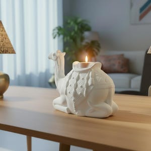 May include: A white ceramic camel-shaped candle holder with a lit candle. The camel is in a seated position with detailed saddle and blanket designs. The candle holder is on a wooden table.