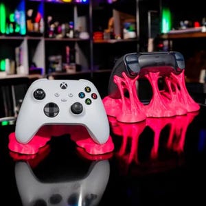 Puede incluir: Dos mandos de juego, uno blanco y otro negro, descansan sobre soportes de color rosa vibrante. El mando blanco, con el logotipo de Xbox, está sobre un soporte con efecto brillante. El mando negro está sobre un soporte similar. Los soportes tienen un diseño artístico único.