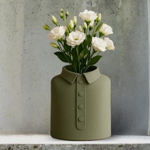 Może przedstawiać: Oliwkowo-zielony ceramiczny wazon w kształcie koszuli, z kołnierzykiem i detalami guzików. Zawiera bukiet białych kwiatów i zielonych liści. Ten nowoczesny design jest idealny do dodania artystycznego akcentu do każdego domu.