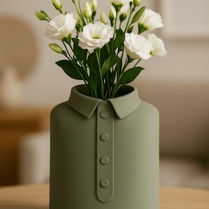 Könnte beinhalten: Eine olivgrüne Keramikvase in Hemdform mit Kragen und Knöpfen. Sie enthält einen Strauß weißer Blumen und grünes Laub. Dieses moderne Design eignet sich ideal, um jedem Zuhause eine künstlerische Note zu verleihen.