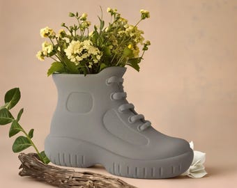 May include: A gray boot-shaped vase filled with yellow flowers. The vase has a lace-up design and a textured side panel. A piece of driftwood and a white flower are in the foreground, with a soft pink background.