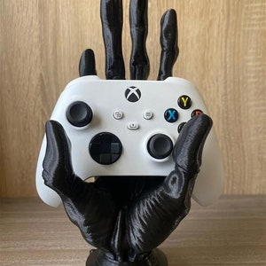 May include: A white Xbox controller held by a black hand-shaped stand. The controller features coloured buttons and the Xbox logo. The stand has a detailed, textured design.