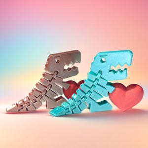 Könnte beinhalten: Zwei 3D-gedruckte Dinosaurierskelettfiguren, eine bronzefarbene und eine türkisfarbene, werden mit zwei roten Herzformen dargestellt. Die Figuren stehen vor einem weichen, farbenfrohen Hintergrund, was ein verspieltes und romantisches Thema erzeugt.