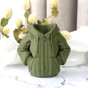 Peut inclure: Un vase en c&eacute;ramique vert olive en forme de veste &agrave; capuche. Le vase pr&eacute;sente une fermeture &eacute;clair sur le devant, des poignets c&ocirc;tel&eacute;s et des poches. Il est pos&eacute; sur une surface en marbre blanc.