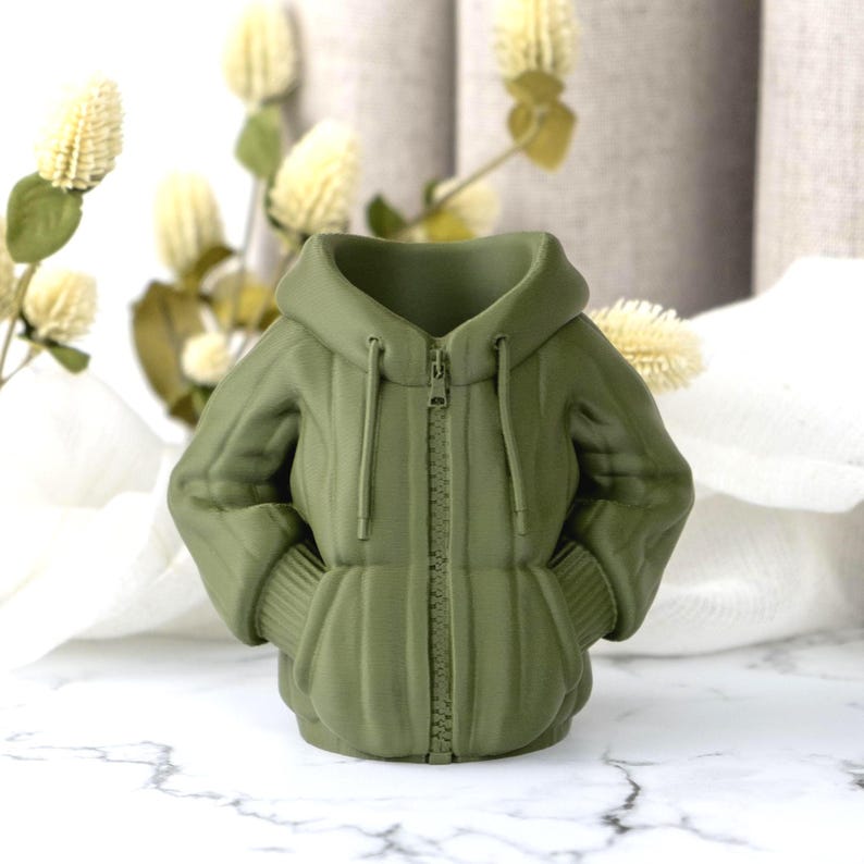 Peut inclure: Un vase en c&eacute;ramique vert olive en forme de veste &agrave; capuche. Le vase pr&eacute;sente une fermeture &eacute;clair sur le devant, des poignets c&ocirc;tel&eacute;s et des poches. Il est pos&eacute; sur une surface en marbre blanc.