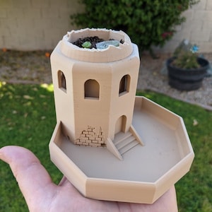 May include: A tan-coloured, 3D-printed miniature castle planter with a circular top and rectangular tower. The tower has arched windows and a small staircase. The planter is filled with soil and a small succulent plant.