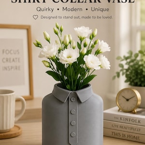Pode incluir: Um vaso de cerâmica cinza fosco em formato de colarinho de camisa com uma carcela central com quatro botões circulares. O vaso contém um buquê de flores brancas com caules e botões verdes. O texto acima diz SHIRT COLLAR VASE.