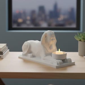 K&ouml;nnte beinhalten: Eine wei&szlig;e Sphinx-Statue aus Keramik mit integriertem Kerzenhalter. Die Statue steht auf einem rechteckigen Sockel und h&auml;lt eine brennende Teelichtkerze. Im Hintergrund eine verschwommene Stadtlandschaft und eine Sukkulentenpflanze.