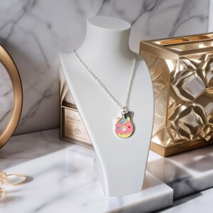 May include: A silver necklace with a pendant shaped like a pink donut with colorful sprinkles and a unicorn horn. The necklace is displayed on a white bust against a marble background. A gold-colored decorative box is in the background.