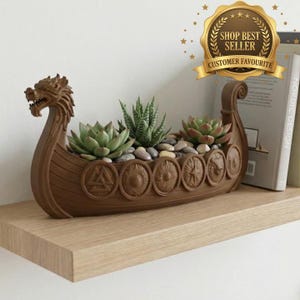 May include: A brown, boat-shaped planter featuring a dragon head at the bow and circular shield motifs along the side. The vessel contains three green succulent plants nestled among small, smooth gray stones, creating a decorative display.