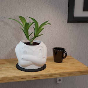 Op de afbeelding: Een witte plantenbak met doktersthema met een groene plant erin, op een houten plank. De plantenbak heeft gekruiste armen en een stethoscoopontwerp. Een zwarte koffiemok staat ernaast.
