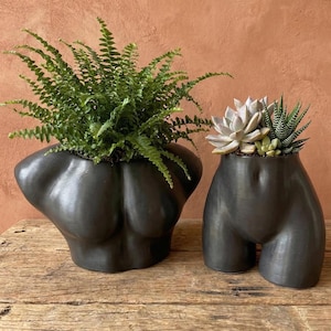 May include: Two matte black ceramic planters shaped like human torsos. The larger planter on the left holds a lush green fern, while the smaller planter on the right contains a cluster of light-coloured succulents and a spiky green plant.