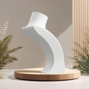 May include: A white, curved jewelry display stand. The stand is on a round, wooden platform. The background features neutral tones and decorative plants. The stand is designed to showcase necklaces or other jewelry.