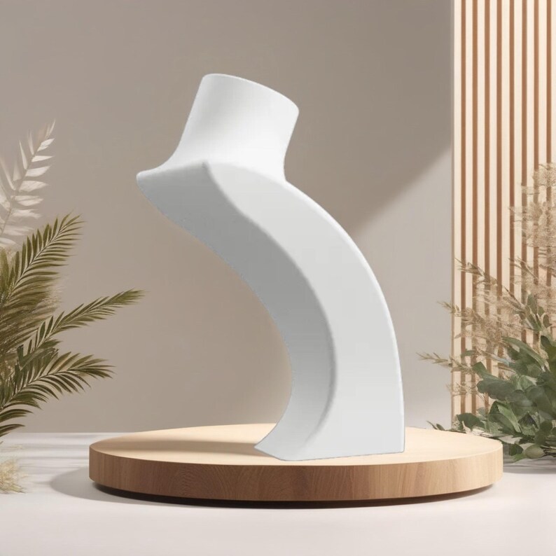 May include: A white, curved jewelry display stand. The stand is on a round, wooden platform. The background features neutral tones and decorative plants. The stand is designed to showcase necklaces or other jewelry.