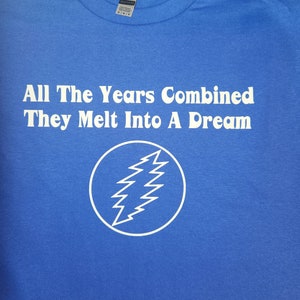 May include: A royal blue t-shirt with white text that reads "All The Years Combined They Melt Into A Dream". A white lightning bolt symbol is centered below the text.