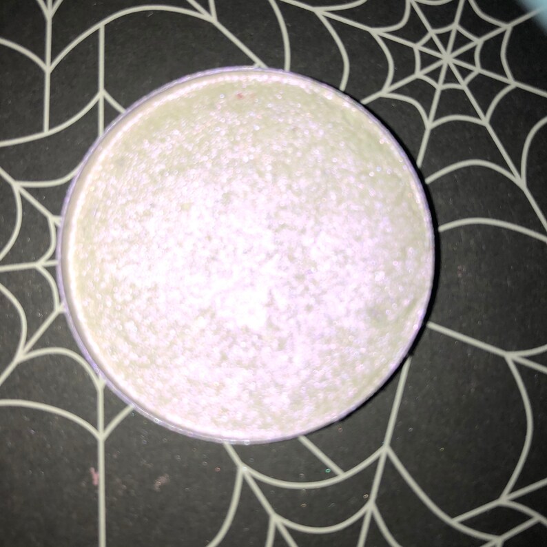 K&ouml;nnte beinhalten: Ein rundes, hellgr&uuml;nes Kosmetikprodukt mit schimmerndem, perlmuttartigem Finish. Das Produkt befindet sich in einem runden Beh&auml;lter mit einem hellvioletten Rand. Der Hintergrund ist schwarz mit einem wei&szlig;en Spinnennetz-Design.