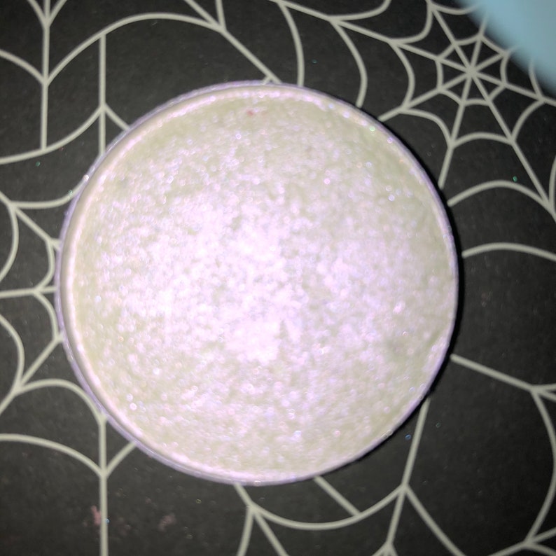 K&ouml;nnte beinhalten: Ein runder, perlmuttartiger Lidschatten-Kompakt mit einem hellgr&uuml;nen Farbton. Der Lidschatten hat eine schimmernde, strukturierte Oberfl&auml;che und befindet sich in einem silberfarbenen Metallgeh&auml;use. Der Kompakt befindet sich vor einem schwarzen Hintergrund mit einem wei&szlig;en Spinnennetz-Design.