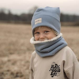 Könnte beinhalten: Ein Kind trägt eine hellblaue Rippstrickmütze und einen passenden Rundschal mit weißem Kunstfellfutter. Das Kind trägt auch einen beigefarbenen Sweatshirt mit dem Text "HAVE A GOOD DAY" und einem Smiley-Motiv.