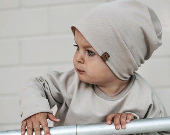 May include: A light beige beanie hat with a small brown rectangular tag on the side, paired with a matching long-sleeved shirt in the same solid light beige color. The beanie features a soft, slouchy fit that covers the head and ears comfortably.