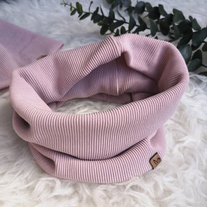 Könnte beinhalten: Ein weicher, rosa gerippter Halsschmeichler oder Schal. Der Halsschmeichler ist auf sich selbst gefaltet und zeigt die Innenseite des Stoffes. Der Halsschmeichler hat ein kleines braunes Leder-Etikett mit dem Buchstaben "M" darauf.