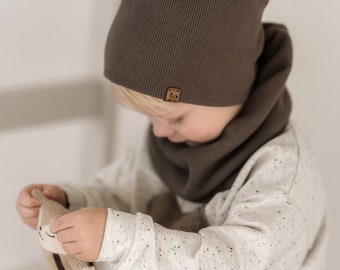 May include: A child wearing a brown beanie and matching neck warmer, holding a knitted toy rabbit. The beanie has a small brown leather patch with a logo on it.