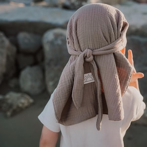 Könnte beinhalten: Ein taupefarbenes Kopftuch aus weichem, strukturiertem Musselin-Stoff. Die Kopfbedeckung verfügt über ein Bindedesign mit langen, losen Enden und ein kleines, weißes, rechteckiges Etikett an der Seite mit einer schwarzen Strichzeichnung einer Sonne.
