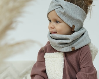 May include: A young child wearing a gray ribbed knit headband and matching neck warmer. The child is wearing a maroon sweater with a white fleece patch pocket.