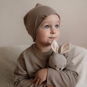 Könnte beinhalten: Ein kleines Kind trägt eine braune Mütze mit einem kleinen braunen Etikett und hält ein graues Stofftier-Kaninchen mit beigefarbenen Ohren. Das Kind trägt ein braunes Langarmshirt.
