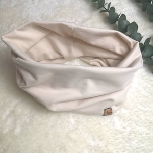 Könnte beinhalten: Ein cremefarbener, weicher und dehnbarer Stoff-Halswärmer mit einem kleinen braunen Lederanhänger.