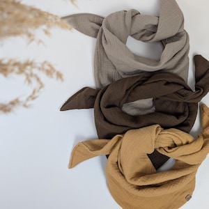 Könnte beinhalten: Drei Baumwoll-Musselin-Quadrate in Beige, Braun und Senfgelb. Die Quadrate sind gefaltet und übereinander gestapelt.