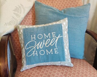 May include: A pink armchair with a floral pattern upholstery. Two throw pillows are on the chair, one is blue and the other is white with the text "Home Sweet Home" in black.