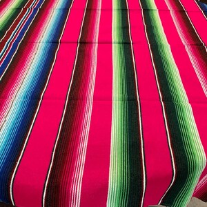May include: A vibrant pink and blue striped Mexican blanket with a traditional design. The blanket is made of soft, woven fabric and features a variety of colors, including pink, blue, green, white, and black.