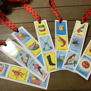 May include: A set of ten colorful bookmarks featuring images and Spanish text from the popular Mexican card game Loteria. The bookmarks are printed on white cardstock and have red and black polka dot ribbon attached.