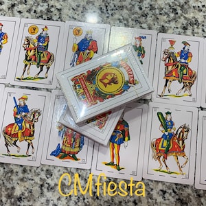 May include: A deck of Spanish playing cards, called "Cartas Españolas", with a white background and colorful illustrations. The cards are arranged in a fan shape, with the box of cards in the center. The box is white with a gold seal and the words "Cartas Españolas" printed on it.