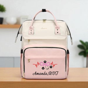 May include: A cream and pink backpack with a top handle and adjustable straps. The front pocket features a pink background with a red heartbeat line, a bow, paw prints, and the text "Amanda, RVT" in black.