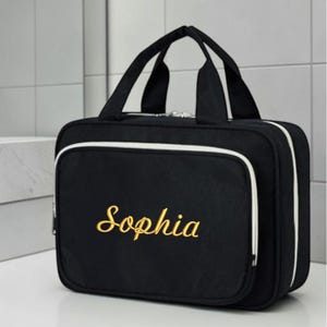 May include: A black rectangular travel toiletry bag with two top handles and a white piped trim. The front features the name Sophia embroidered in gold cursive script. The bag has a zippered main compartment and a smaller front zippered pocket.